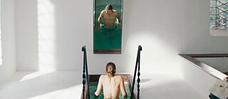 Scene from the film “Skin Deep”. A person entering a narrow indoor pool via small stairs; a vertical wall mirror reflects the upper body in the water.