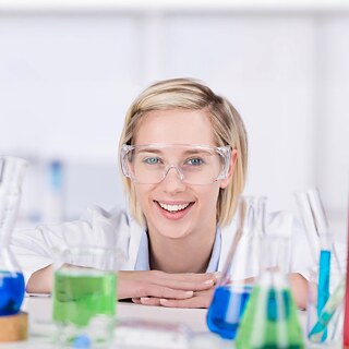 Person mit Schutzbrille vor Glasgefäßen mit bunten Flüssigkeiten.