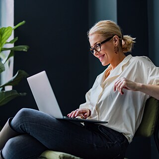 Eine Frau arbeitet am Laptop. Sie sitzt entspannt auf einem Sessel und nimmt an einem Videocall teil. Porträtaufnahme.