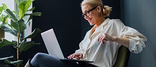 Eine Frau arbeitet am Laptop. Sie sitzt entspannt auf einem Sessel und nimmt an einem Videocall teil. Porträtaufnahme.