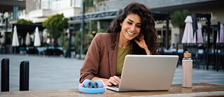 Eine Frau arbeitet im Freien am Laptop. Sie lächelt zufrieden. Porträtaufnahme.