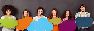 Eine Gruppe Männer und Frauen stehen nebeneinander vor einer dunklen Wand und halten große, bunte Sprechblasen vor sich. Sie haben Spaß und lachen. in die Kamera. Textfreiraum