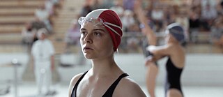 Scene from the film “Beyond the Blue Border”. A person wearing a red swim cap and goggles stands by the edge of an indoor pool; other swimmers and spectators are blurred in the background.