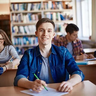 Person schreibt im Klassenraum, weitere Personen im Hintergrund.