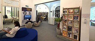Bibliothek mit Sitzmöbeln; mehrere Personen lesen oder nutzen eine Gaming-Station, im Hintergrund sind Bücherregale zu sehen.