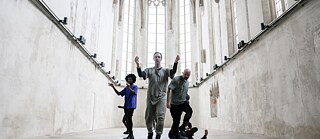 In der Mitte eines leeren Kirchenraumes laufen drei männliche Tänzer in unterschiedlichen Posen auf die Kamera zu.