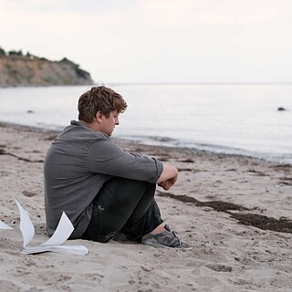 Scene from the film “Afire”. A person sitting on a beach with knees drawn up, facing the sea; loose pages lie on the sand nearby, with a coastline in the background.