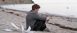 Scene from the film “Afire”. A person sitting on a beach with knees drawn up, facing the sea; loose pages lie on the sand nearby, with a coastline in the background.