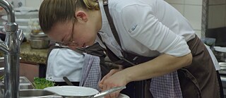 In einer professionellen Küche beugt sich eine junge Frau über eine Gefäß sich auf die Zubereitung einer Speise konzentrierend