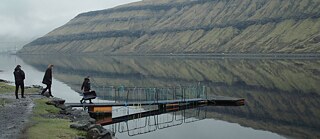 Panorama einer Fjordlandschaft. Im der mittleren Distanz gehen drei Personen auf einen Steg zu, der ins Wasser führt.