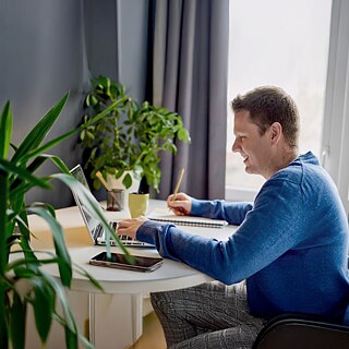 Person schreibt am Tisch neben Laptop, Pflanzen und Vorhänge im Hintergrund.