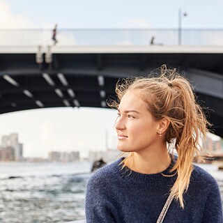 Eine junge Deutschlehrerin sitzt an einem Fluss und blickt nachdenklich in die Ferne.​