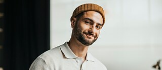 Ein junger Deutschlehrer steht in seinem Klassenzimmer vor einer digitalen Tafel und blickt freundlich lächelnd in die Kamera.​