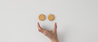 Ein lachender Smiley mit zwei Keksen als Augen und einer geöffneten Hand als Mund
