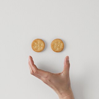 Ein lachender Smiley mit zwei Keksen als Augen und einer geöffneten Hand als Mund