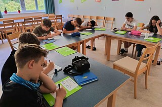 In einem Klassenzimmer sitzen Jugendliche an in U-Form angerichteten Tischen und schreiben.