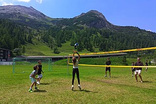 Jugendliche spielen Volleyball auf einer Wiese. Im Hintergrund sind hohe, z.T. bewaldete Berge zu sehen.