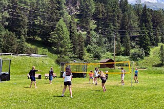 Jugendliche spielen Volleyball auf einer Wiese. im Hintergrund sind ein Wald und Berge zu sehen.