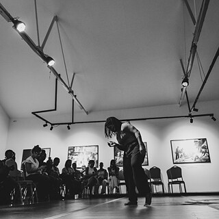 Schwarz-Weiß-Fotografie einer Tanzperformance in einem Ausstellungsraum. Im Vordergrund bewegt sich eine Person auf einer freien Fläche, während das Publikum auf Stühlen am Rand sitzt. An den Wänden hängen gerahmte Fotografien, und Deckenstrahler beleuchten den Raum.