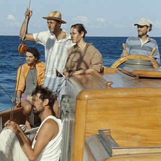 Filmszene aus „Human Flowers of Flesh“: Fünf Personen auf dem Segelschiff im Meer, eine steuert, andere stehen und sitzen an Deck