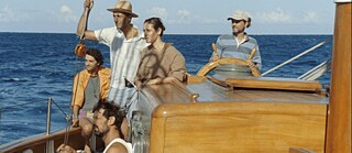 Filmszene aus „Human Flowers of Flesh“: Fünf Personen auf dem Segelschiff im Meer, eine steuert, andere stehen und sitzen an Deck