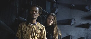 Zwei Personen (Mukenge und Schellhammer) vor einer dunklen Kunstinstallation mit schwarzen, kegelförmigen Stoffelementen.
