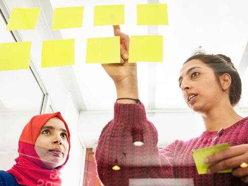 Two people who are perceived as female stand before a glass wall, which is covered with eight yellow sticky notes in a well-lit room.