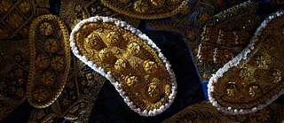 Nahaufnahme mehrerer reich verzierter textile Elemente in Gold‑ und Blautönen. Im Mittelpunkt liegen längliche, ovale Ornamente mit erhabenen kreisförmigen Mustern, goldenen Stickereien und weißen Perlen entlang der Ränder. Umgeben sind sie von dicht bestickten Stoffflächen mit floralen und geometrischen Motiven. Das Bild zeigt feine Texturen, metallische Oberflächen und dekorative Details auf dunkelblauem Hintergrund.