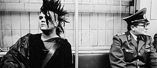 Ein Punk mit Irokesenschnitt und ein uniformierter Volkspolizist warten nebeneinander auf die U-Bahn, dabei sehr bemüht sind, sich gegenseitig zu ignorieren.