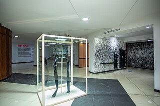 Heller Museumsraum mit einer schwarzen, stachelbesetzten Skulptur in einer Glasvitrine. Im Hintergrund sind eine beschriftete Interaktionszone mit Wandzeichnungen, Texten und textile Kunstobjekte zu sehen.