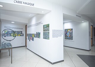 Ausstellungsansicht mit Texttafeln, Bilddokumentationen und einer Vitrine. Die Bereiche „Carré Magique“ und „Partagisme“ präsentieren Fotografien, grafische Arbeiten und erläuternde Wandtexte.