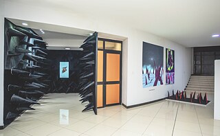 Ein Ausstellungsraum mit einer begehbaren Installation aus schwarzen, kegelförmigen Textilelementen, die eine Wandöffnung umrahmen. Rechts sind zwei großformatige Kunstwerke mit violetten und bläulichen Farbtönen sowie eine bodennahe Skulptur aus schwarzen und roten Spitzen zu sehen.