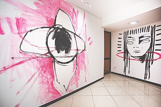 Großformatige Wandzeichnungen in Schwarz und Pink bedecken die Wände eines Galerieraums. Links ist eine abstrahierte Figur mit kreisförmigen Linien dargestellt, rechts ein stilisiertes Gesicht mit vertikalen schwarzen Linien und einem pinken Ring.