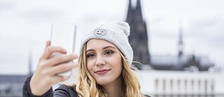 Eine junge Frau mit grauer Mütze fotografiert sich lächelnd mit dem Smartphone. Im Hintergrund der Kölner Dom