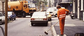 La imagen muestra una diapositiva antigua: la recogida de basuras en Dortmund. En primer plano, una persona vestida de naranja barre una calle. Al fondo, un camión de la basura dobla la esquina.