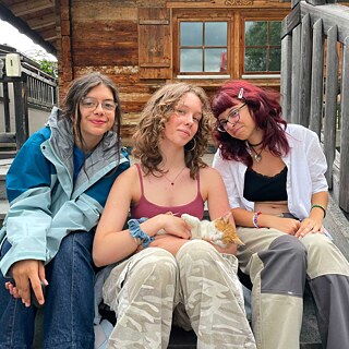 Drei junge Frauen sitzen auf der Holztreppe zu einer Hütte und streicheln eine junge Katze.