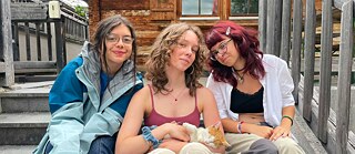 Drei junge Frauen sitzen auf der Holztreppe zu einer Hütte und streicheln eine junge Katze.