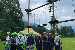 Mit Sicherheitsgurten und Helmen ausgestattete Jugendliche stehen unter einem Gerüst in eime Kletterpark