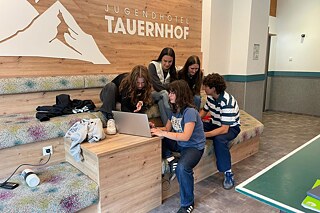 Jugendliche sitzen auf einer Bank und betrachten den Monitor eines Laptops.