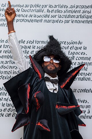 Performance-Kunst mit avantgardistischem Kostüm vor den Ausstellungstexten von „Elima-NoBody“ in der Ausstellung „Elima-NoBody“ im Musée d'Art Contemporain et Multimédia in Kinshasa.