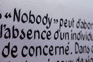 Nahaufnahme eines Ausstellungstextes mit dem hervorgehobenen Wort „Nobody“ auf einer weißen Wand in der Ausstellung „Elima-NoBody“ im Musée d'Art Contemporain et Multimédia in Kinshasa.