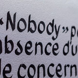 Nahaufnahme eines Ausstellungstextes mit dem hervorgehobenen Wort „Nobody“ auf einer weißen Wand in der Ausstellung „Elima-NoBody“ im Musée d'Art Contemporain et Multimédia in Kinshasa.