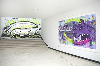 Heller Ausstellungsraum mit einer Treppe und zwei großformatigen Kunstwerken. Links ist ein abstraktes Motiv in Schwarz, Weiß und Neongrün zu sehen, rechts ein ausdrucksstarkes violettes Wesen mit leuchtenden gelben und pinken Akzenten.