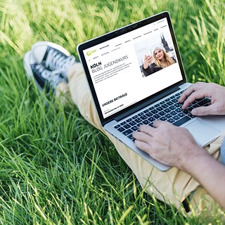 Mann sitzt mit ausgestreckten Beinen auf dem Rasen und tippt in ein Laptop.