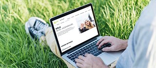 Mann sitzt mit ausgestreckten Beinen auf dem Rasen und tippt in ein Laptop.