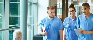 Drei medizinische Fachkräfte in blauer Arbeitskleidung besprechen Informationen auf einem Tablet in einem modernen Klinikflur.
