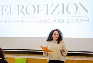 A person stands in front of a large projection screen displaying the text “EVROVIZION.CROSSING STORIES AND SPACES” holding an orange sheet of paper and a smartphone. The image depicts a presentation or event in an indoor venue.