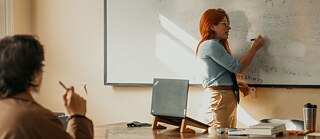 Eine Frau schreibt am Whiteboard vor einer Klasse, in der eine Person in der ersten Reihe sitzt.