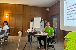 Ein junger Erwachsener sitzt an einem Tisch und spricht in ein Microphon.