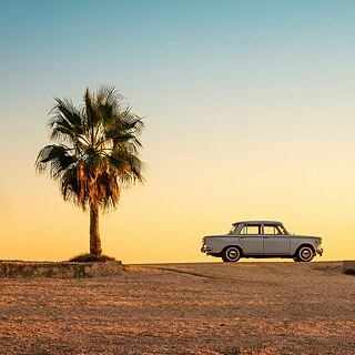 Klassisches Auto zwischen zwei Palmen vor einem goldenen Sonnenuntergang.